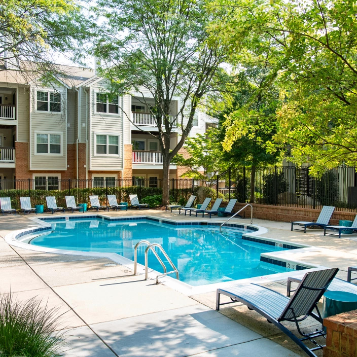 Community pool area surrounded by lush trees, lounge chairs, and multi-story residential buildings