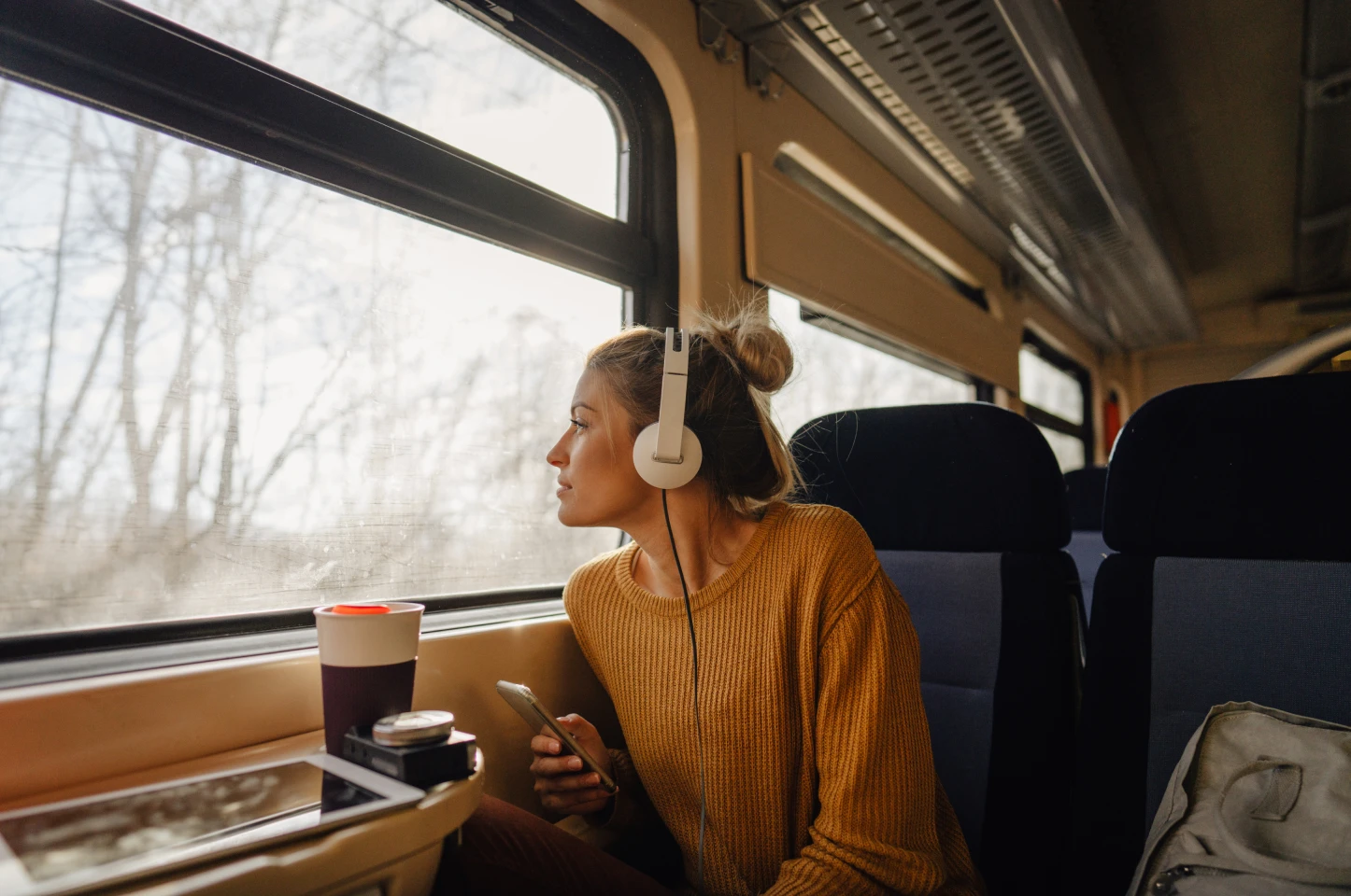 Woman wearing headphones gazing out train window while listening to music and holding phone